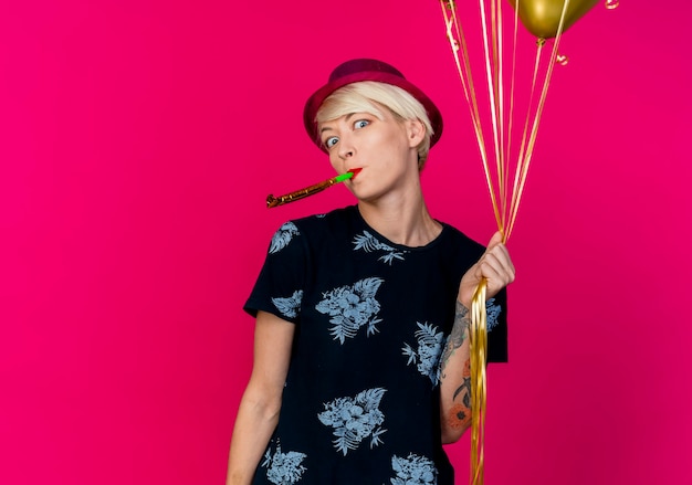 Kostenlose Foto beeindrucktes junges blondes partygirl, das partyhut hält, der ballons hält, die kamera betrachten und partygebläse bläst, das auf purpurrotem hintergrund mit kopienraum lokalisiert wird