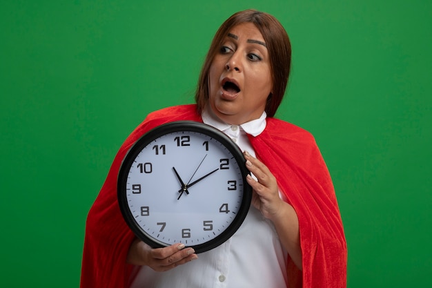 Kostenlose Foto erschrockenes superheldenweibchen mittleren alters, das seite betrachtet, die wanduhr lokalisiert auf grün hält