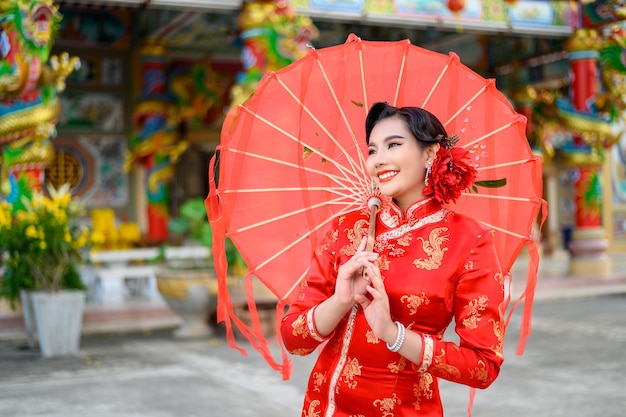 Gratis Bild porträtt av en vacker asiatisk kvinna som bär en cheongsam, ler och poserar med ett rött pappersparaply vid en helgedom under kinesiskt nyår